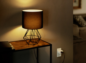  The Eve Energy plug, inserted into a standard wall outlet with green LED power indicator ON, connected to a lamp.