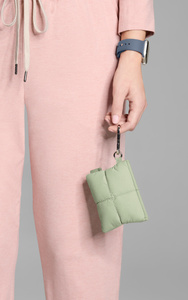 Comfyable Puffer AirPods Case, side exterior, held in a person’s left by chrome carabiner attached at side