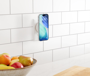 Twelve South PowerBug Qi2.2-oplader in keukenstopcontact, een hand brengt een iPhone naar het laadoppervlak, doorgesneden peer en snijplank voor een fruitschaal op de achtergrond.