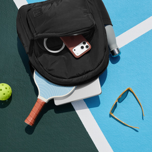 Herschel Cloudform Backpack XL, front exterior, black color, lying on striped court surface. Front pouch zipper open, iPhone inserted, top zipper pouch open. iPad and pickleball racquet inserted, sunglasses and water bottle
