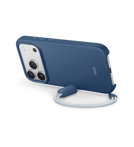 Coque Beats Kickstand Bleu basalte protégeant un iPhone 17 Pro, vu de dos en mode paysage, l’adaptateur magnétique de la dragonne est fixé sur le côté de la coque et fait tenir l’iPhone en position verticale sur un bureau en bois