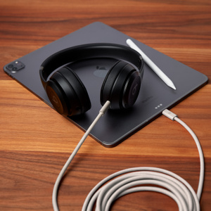Beats USB-C to USB-C Woven Cable in Surge Stone plugged into Beats Solo 4 headphones and iPad Pro, coiled up to show 3 m / 10 ft length. 