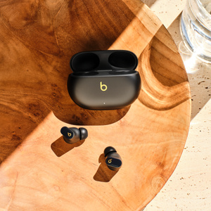 Fones de ouvido Beats Studio Buds+ e estojo de recarga em preto e dourado sobre uma mesa, ao lado de um copo de água 