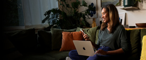 A person sitting on a couch with a silver MacBook Air in their lap, legs crossed, smiling while holding a silver iPhone