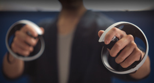A person holds PlayStation VR2 Sense Controllers, one in each hand, white and black two-tone orb-shaped body, fingers on the trigger