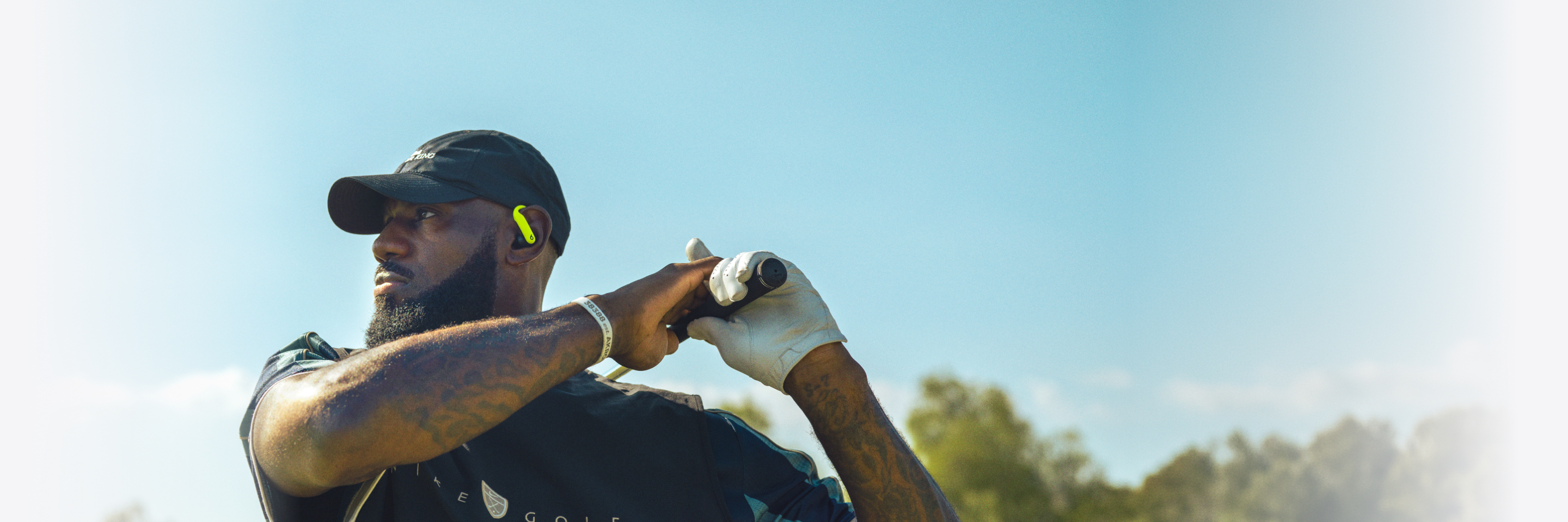 LeBron James 佩戴着黑配荧光黄色 Nike 特别版 Powerbeats Pro 2 耳机，手持高尔夫球杆，左侧耳机上印有黑色 Nike 标志。