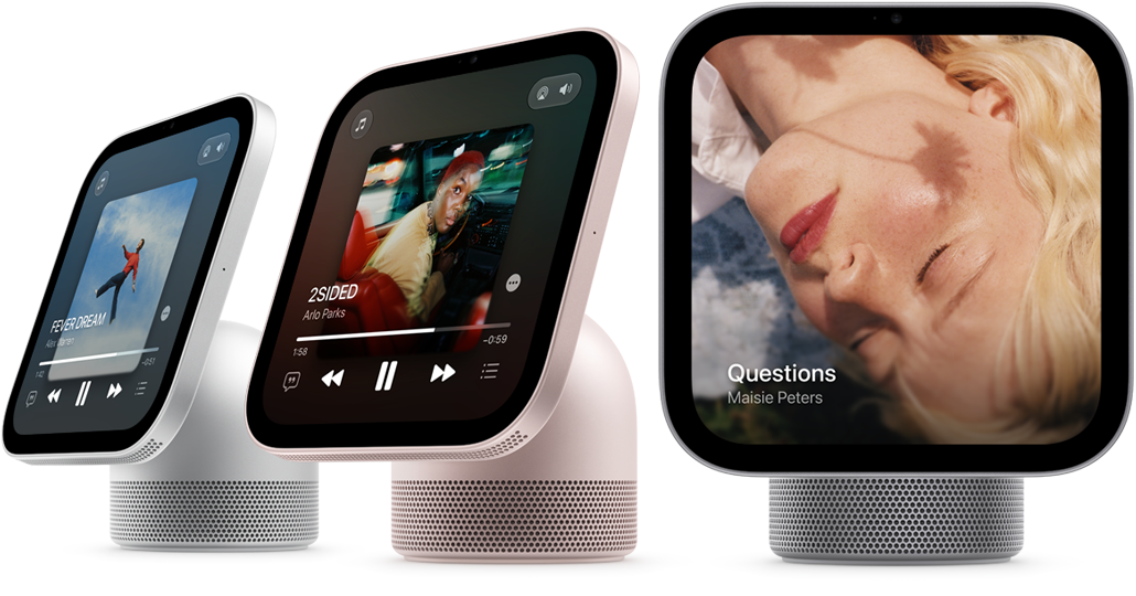 Apple HomeView, display with base, colour-matching domed cylindrical speaker base with circular perforations. Three devices: silver colour, screen displays Apple Music Now Playing, album artwork, playback controls. Rose pink colour, screen displays Apple Music Now Playing, album artwork, playback controls. Silver colour, screen displays album cover and song by Maisie Peters