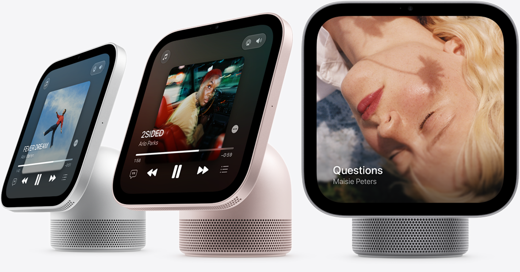 Apple HomeView, display with base, color-matching domed cylindrical speaker base with circular perforations. Three devices: silver color, screen displays Apple Music Now Playing, album artwork, playback controls. Rose pink color, screen displays Apple Music Now Playing, album artwork, playback controls. Silver color, screen displays album album cover and song by Maisie Peters