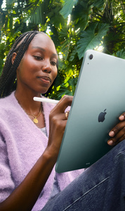 A person sits outdoors among tropical foliage, holding blue iPad Air and Apple Pencil in hand
