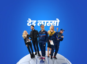 Apple TV+ promo, title: Ted Lasso, Jason Sudeikis smiling broadly, Juno Temple smiling with her hand on her hip, Brett Goldstein in a leather jacket, Hannah Waddingham with fist raised, Brendan Hunt holding a clipboard, cast fanned out on an oversized soccer ball, blue gradient background.